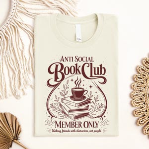 May include: A cream-coloured t-shirt featuring a dark brown graphic of a stack of three books topped with a steaming cup of coffee. The text reads Anti Social Book Club Member Only, Making friends with characters, not people, in a decorative font.