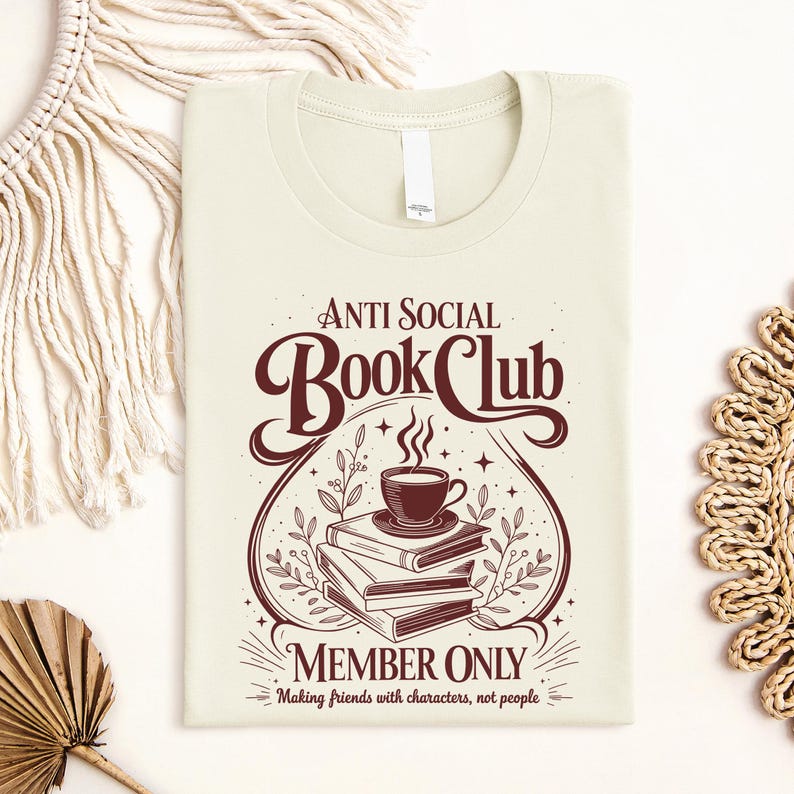 May include: A cream-coloured t-shirt featuring a dark brown graphic of a stack of three books topped with a steaming cup of coffee. The text reads Anti Social Book Club Member Only, Making friends with characters, not people, in a decorative font.