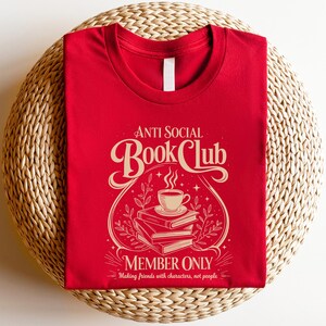 May include: A red crew neck t-shirt featuring a cream-coloured graphic with the text Anti Social Book Club Member Only Making friends with characters, not people. The design includes a stack of three books topped with a steaming cup of coffee.