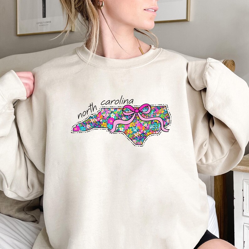 May include: A beige crewneck sweatshirt featuring a graphic of the state of North Carolina filled with a colorful floral pattern and accented by a bright pink ribbon bow. The text north carolina is printed in black above the state shape.
