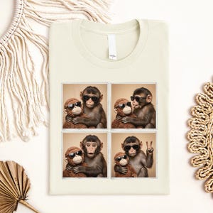 May include: A cream-coloured t-shirt featuring a graphic print of four square photo frames. Each frame shows a monkey wearing black sunglasses and hugging a brown plush monkey toy. The monkey poses differently in each frame, including a peace sign.