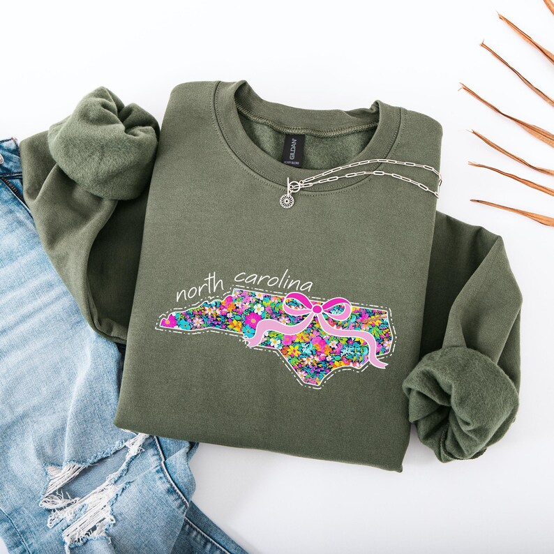 May include: An olive green crewneck sweatshirt featuring a graphic of the state of North Carolina filled with a colorful floral pattern and accented with a pink ribbon. The text north carolina is printed in white above the state outline.