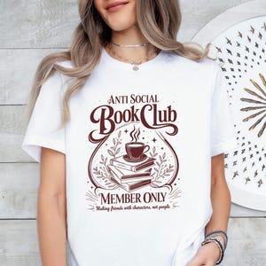 May include: A white t-shirt featuring a dark brown graphic design of a stack of books topped with a steaming cup of coffee. The text reads Anti Social Book Club Member Only Making friends with characters, not people, surrounded by botanical accents.