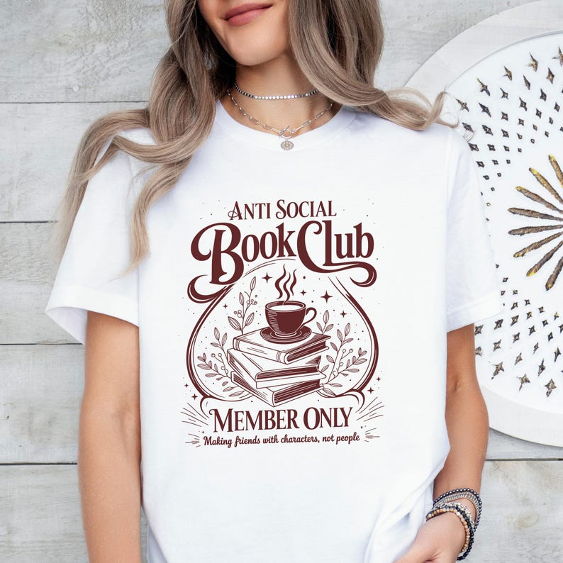 May include: A white t-shirt featuring a dark brown graphic design of a stack of books topped with a steaming cup of coffee. The text reads Anti Social Book Club Member Only Making friends with characters, not people, surrounded by botanical accents.