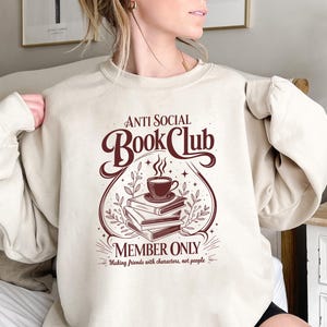 May include: A beige crewneck sweatshirt featuring a dark brown graphic print. The design displays a stack of three books topped with a steaming cup of coffee, surrounded by foliage and the text "ANTI SOCIAL BOOK CLUB MEMBER ONLY Making friends with characters, not people."