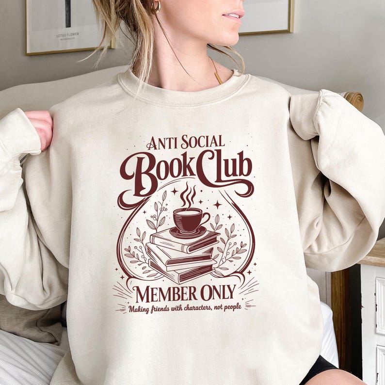 May include: A beige crewneck sweatshirt featuring a dark brown graphic print. The design displays a stack of three books topped with a steaming cup of coffee, surrounded by foliage and the text "ANTI SOCIAL BOOK CLUB MEMBER ONLY Making friends with characters, not people."