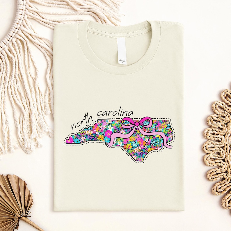 May include: A cream-colored t-shirt featuring a graphic of the state of North Carolina filled with a colorful floral pattern. A bright pink ribbon bow is tied around the center of the state shape. The text north carolina is printed in black.