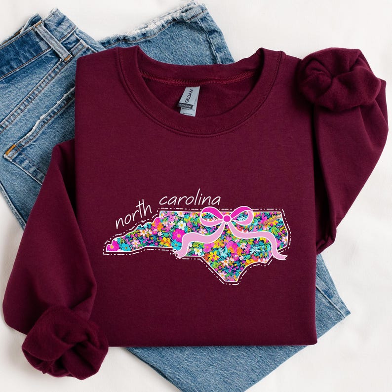 May include: A maroon long-sleeve sweatshirt featuring a graphic of the state of North Carolina filled with a colorful floral pattern. A pink ribbon bow is placed over the state graphic, and the text north carolina is written in white script.