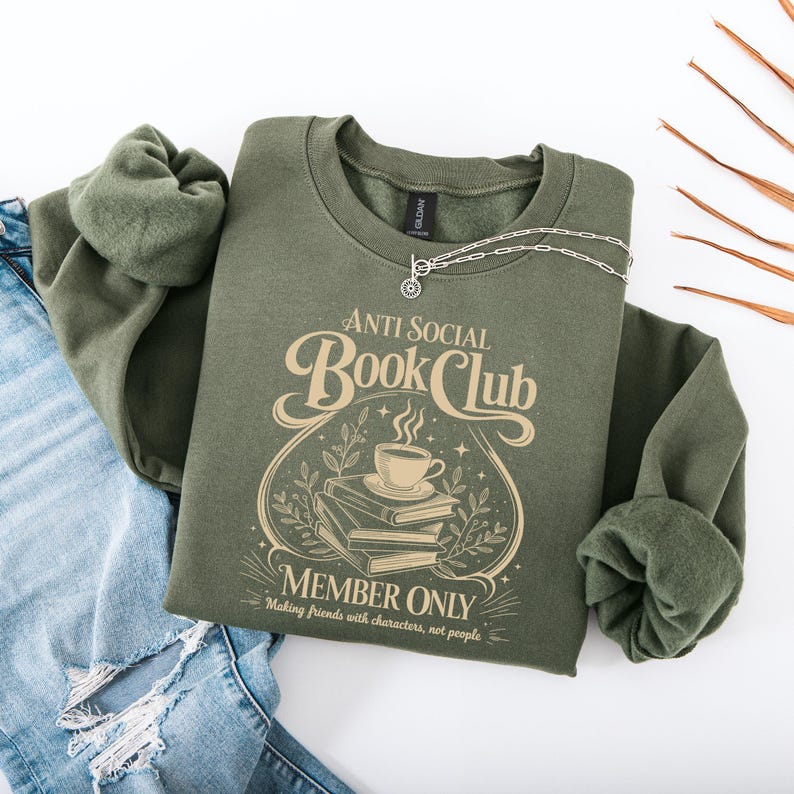 May include: An olive green crewneck sweatshirt featuring a cream-coloured graphic with the text "Anti Social Book Club Member Only Making friends with characters, not people" surrounding an illustration of a steaming coffee cup atop a stack of books.