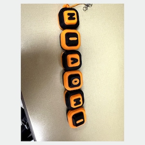 May include: A vertical keychain featuring six square, rounded-edge blocks arranged in a column. Each block has a black center with an orange border and displays a single embossed letter in orange. The letters spell out N-I-A-O-M-I from top to bottom.