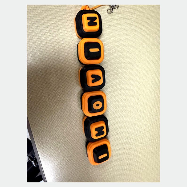 May include: A vertical keychain featuring six square, rounded-edge blocks arranged in a column. Each block has a black center with an orange border and displays a single embossed letter in orange. The letters spell out N-I-A-O-M-I from top to bottom.