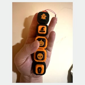 May include: A vertical string of five square, alternating black and orange silicone beads featuring embossed Halloween icons including a ghost, a witch hat, a jack-o'-lantern, a skull, and a coffin, attached to a thin pink cord loop.