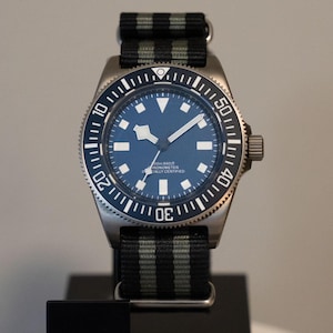 May include: A stainless steel wristwatch with a dark blue dial, white geometric hour markers, and a rotating bezel with white numerals. The watch features a black and grey striped nylon strap with silver-toned metal hardware and a crown on the side.