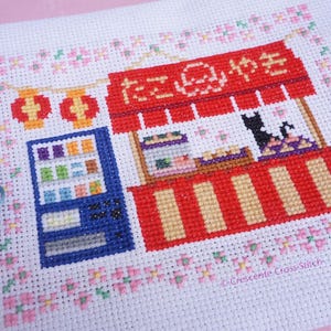 May include: A cross-stitch embroidery on white fabric featuring a red takoyaki food stall with Japanese text, a blue vending machine, two red lanterns, and a black cat. The design is framed by a border of small pink and green floral motifs.