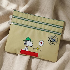 May include: A light olive green zippered pouch featuring a graphic of Snoopy as a Beagle Scout wearing a green hat, sitting on a red surface. The pouch includes a circular logo that reads Beagle Scouts 50 years est. 1974 and two silver zippers.
