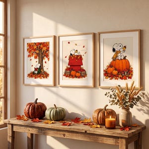 May include: Three framed wall art prints featuring Snoopy in autumn scenes with orange leaves, pumpkins, and a red doghouse. Below, a wooden console table displays three real pumpkins, a lit candle in a glass jar, and a vase with dried wheat.
