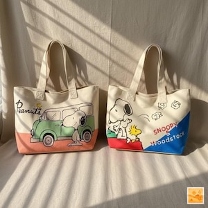Könnte beinhalten: Zwei Tragetaschen aus Canvas mit cremefarbenen Griffen und Körpern. Eine Tasche zeigt Snoopy mit einem grünen Van und dem Wort "Peanuts". Die andere Tasche hat Snoopy und Woodstock mit den Worten "Snoopy & Woodstock" und einer roten und blauen Ecke.