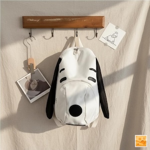 Op de afbeelding: Witte rugzak met zwarte accenten, die lijkt op een cartoonhond. Het heeft een grote zwarte cirkel en gebogen zwarte vormen. De rugzak hangt aan een houten rek met haken. Een kleine tekening en een foto hangen ook.