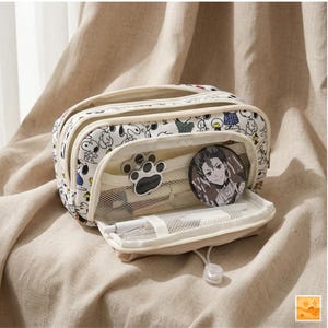 Peut inclure: Trousse à crayons crème avec un imprimé de dessin animé Snoopy. L'étui comporte plusieurs compartiments, dont une poche en filet et une section zippée. Un aimant en forme de patte et un badge rond de personnage sont visibles à l'intérieur.