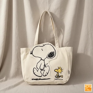 May include: Cream canvas tote bag with a Snoopy and Woodstock design. The bag has two handles and a rectangular shape. The Snoopy illustration is outlined in black with a black ear and Woodstock is yellow.
