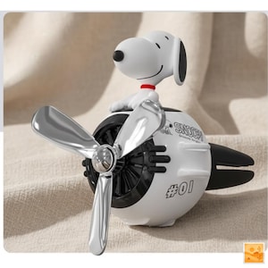 May include: A white Snoopy figure sits atop a white and black airplane-shaped car air freshener. The air freshener has a silver propeller and the text "Snoopy" and "#01" printed on it. The figure has a red collar.
