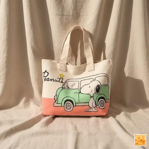 Może przedstawiać: Kremowa torba typu tote z kreskówkową ilustracją Snoopy'ego obok zielonego vana. Torba ma brzoskwiniowe dno i napis "Peanuts" w skrypcie. Ma dwa kremowe uchwyty.