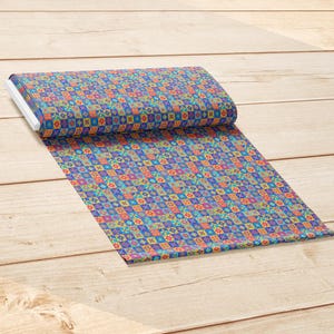 Op de afbeelding: Een rol stof met een herhalend geometrisch patroon van kleine vierkantjes in levendige tinten blauw, oranje, geel, groen en paars. Het textiel is aan &eacute;&eacute;n kant gevouwen en toont een dicht, kleurrijk raster van gedetailleerde stervormige motieven.