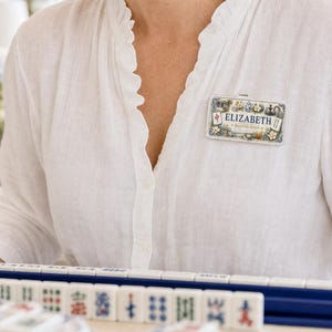 Pode incluir: Um crachá retangular com o nome ELIZABETH em letras azuis, rodeado por uma ilustração colorida de peças de mahjong e motivos florais. O crachá está preso a uma camisa branca, posicionado acima de uma fileira de peças de mahjong.