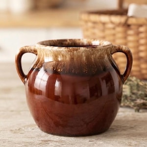 May include: A round ceramic pot with two side handles, featuring a glossy dark brown base that transitions into a lighter cream and tan drip glaze around the upper rim and handles. The vessel has a wide opening and a smooth, reflective surface.