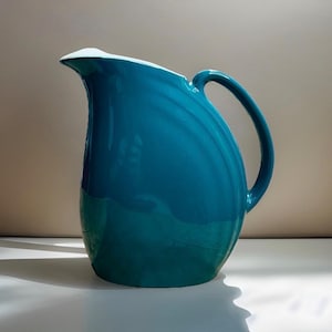 May include: A glossy teal ceramic pitcher featuring a curved silhouette, a wide pouring spout, and a single arched handle. The exterior surface has a smooth, reflective finish with subtle vertical ridges along the side, and the interior is white.