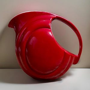 May include: A glossy, bright red ceramic pitcher featuring a distinctive circular, disc-like body with a curved handle that connects from the top rim to the lower side. The vessel has a prominent pouring spout and concentric circular embossed detailing.