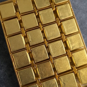 May include: A gold-coloured tray filled with individually wrapped square chocolates. The chocolates are arranged in a grid pattern, each with a shiny, metallic appearance. The tray is also gold. The image is taken from above.
