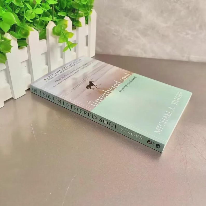 May include: A hardcover book titled "The Untethered Soul" by Michael A. Singer. The cover features a light green and white design with a silhouette of a person. The book is on a reflective surface.