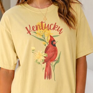 Könnte beinhalten: Ein hellgelbes T-Shirt mit kurzen Ärmeln und einem Grafikdruck eines roten Kardinalvogels, der zwischen gelben Blumen sitzt. Das Wort Kentucky ist in roter Schreibschrift über der Vogelillustration auf der Brust aufgedruckt.
