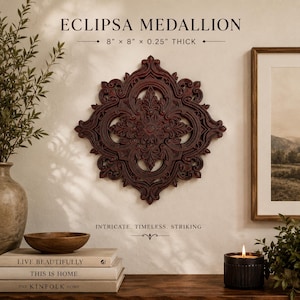 May include: The Eclipsa Medallion is a dark brown, square-shaped wall decor piece with an intricate, symmetrical carved pattern. It measures 8 inches by 8 inches by 0.25 inches thick. The design features ornate, scrolling details and a central floral motif.