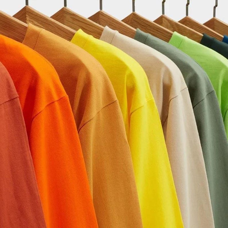 Pode incluir: Uma fila de camisetas penduradas em cabides de madeira, exibidas em um gradiente de cores incluindo terracota, laranja brilhante, amarelo mostarda, amarelo lim&atilde;o, bege, verde s&aacute;lvia, verde lim&atilde;o e verde floresta escuro, todas de manga curta.