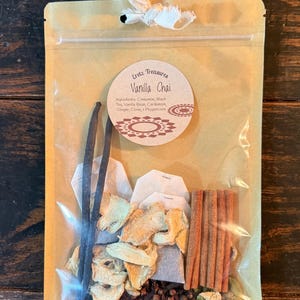 Może przedstawiać: Beżowa papierowa torebka zawierająca składniki herbaty Vanilla Chai, w tym dwie laski wanilii, suszony imbir, cztery laski cynamonu, goździki, ziarna pieprzu, zielony kardamon, trzy torebki herbaty oraz etykietę z napisem Eretz Treasures Vanilla Chai.
