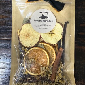 Puede incluir: Una bolsa de plástico transparente con reverso de papel marrón que contiene rodajas de naranja y manzana secas, ramas de canela, vainas de vainilla y flores de manzanilla. Una etiqueta redonda dice Hogwarts Hearthstone con los ingredientes.