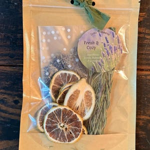 May include: A tan pouch with a green ribbon tie containing dried lemon slices, rosemary sprigs, and lavender buds. A circular label reads Eretz Treasures, Fresh & Cozy, and lists ingredients as Lemon, Rosemary & Lavender with a lavender illustration.