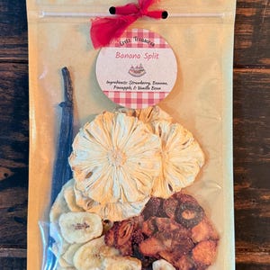 Puede incluir: Una bolsa de plástico transparente con rodajas de fruta deshidratada que incluyen piña, plátano y fresa, además de una vaina de vainilla. Una etiqueta redonda dice Eretz Treasures Banana Split. Un lazo de tela roja está atado en la parte superior.