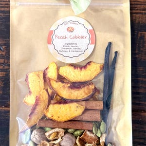 May include: A clear pouch labeled Peach Cobbler containing dried peach slices, cinnamon sticks, whole nutmeg, green cardamom pods, and a vanilla bean. The bag is sealed with a green and coral fabric ribbon tied at the top center of the package.