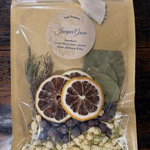 Op de afbeelding: Een doorzichtig plastic zakje met gedroogde botanische ingrediënten, waaronder twee schijfjes citroen, jeneverbessen, jasmijnbloemen, rozemarijntakjes en laurierblaadjes. Een rond etiket vermeldt Eretz Treasures, Juniper Grove, Ingredients: Juniper Berry, Lemon, Jasmine Flower, Rosemary, & Bay.