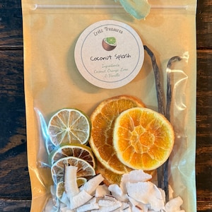 Könnte beinhalten: Ein beigefarbener Beutel mit grünem Band enthält getrocknete Früchte und Kokosnuss. Ein rundes Etikett zeigt Eretz Treasures Coconut Splash mit den Zutaten Coconut, Orange, Lime, & Vanilla. Enthalten sind getrocknete Limettenscheiben, Orangenscheiben, Vanilleschoten und Kokosflocken.