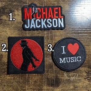 May include: Three embroidered patches. Patch 1 features the text MICHAEL JACKSON in red and white with a silhouette figure. Patch 2 is a square with a red circle and a black silhouette figure. Patch 3 is a circle with the text I HEART MUSIC.