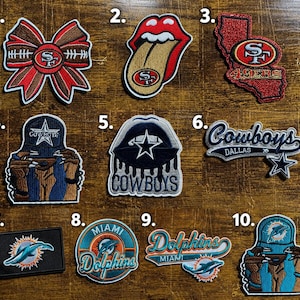 May include: A collection of ten embroidered patches featuring NFL team logos and designs. The patches include San Francisco 49ers bows, lips, and state shapes, Dallas Cowboys hats, lips, and script logos, and Miami Dolphins rectangles, circles, and hats.