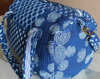May include: A cylindrical quilted duffel bag featuring a deep blue and white fabric with various patterns including concentric circles, dots, and floral motifs. The bag includes a matching fabric shoulder strap attached with brass-toned metal hardware.