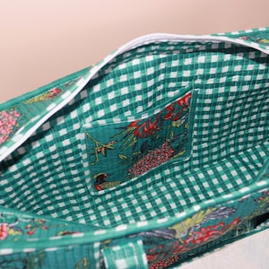 May include: A quilted fabric tote bag featuring a green and white gingham interior lining with a matching open pocket. The exterior displays a vibrant floral pattern in shades of green, red, and yellow, finished with a white zipper closure.
