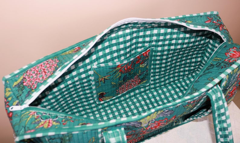 May include: A quilted fabric tote bag featuring a green and white gingham interior lining with a matching open pocket. The exterior displays a vibrant floral pattern in shades of green, red, and yellow, finished with a white zipper closure.