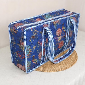 May include: A rectangular quilted bag in deep blue featuring a vibrant floral and bird pattern in red, orange, and green. The bag is finished with white and blue striped fabric trim along the edges and includes two matching striped handles.