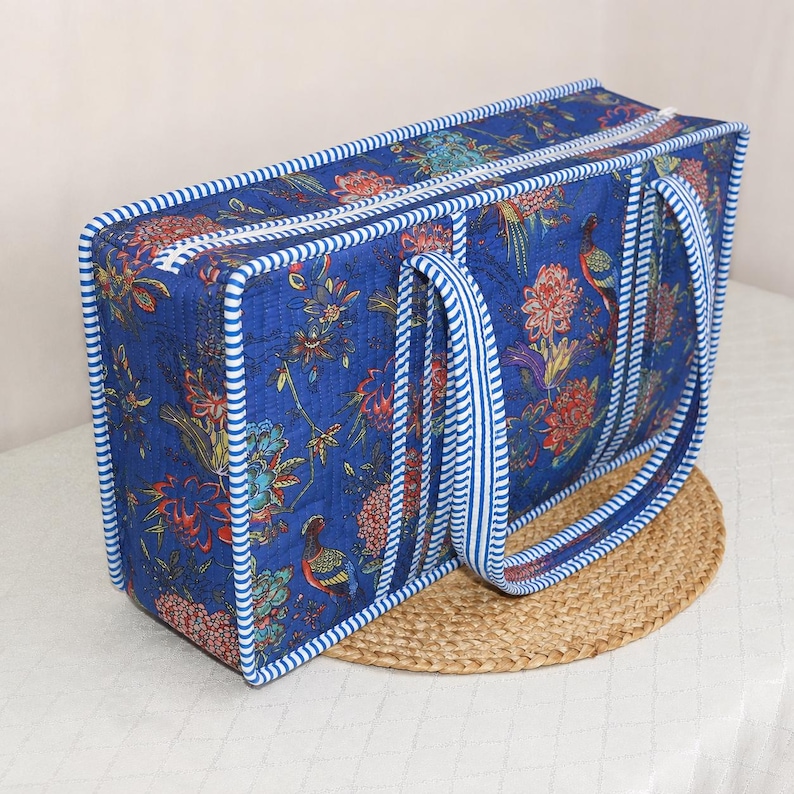 May include: A rectangular quilted bag in deep blue featuring a vibrant floral and bird pattern in red, orange, and green. The bag is finished with white and blue striped fabric trim along the edges and includes two matching striped handles.